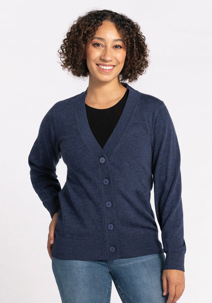 A smiling person with curly hair is wearing the Gia Sweater - Twilight Storm from Woolx over a black shirt and light blue jeans, standing against a plain white background. 