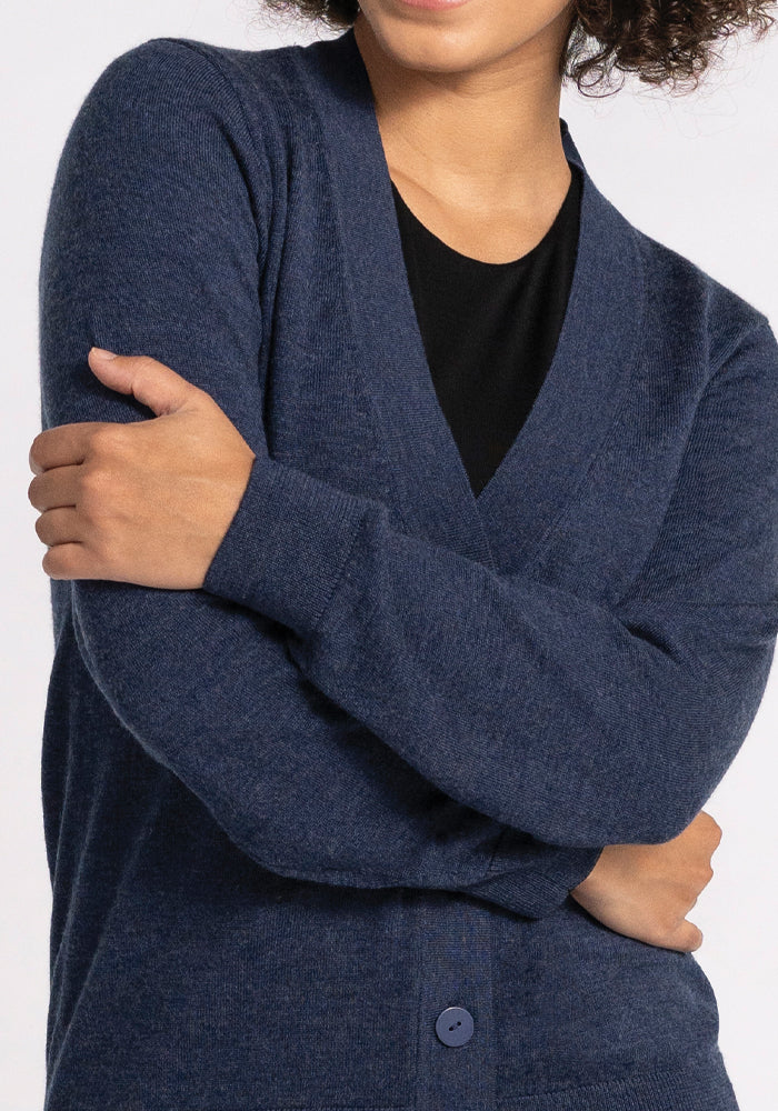 A person is seen wearing the Gia Sweater in Twilight Storm, a dark blue cardigan from Woolx. Their arms are crossed, and the cardigan is fully buttoned. The backdrop is plain and light-colored.