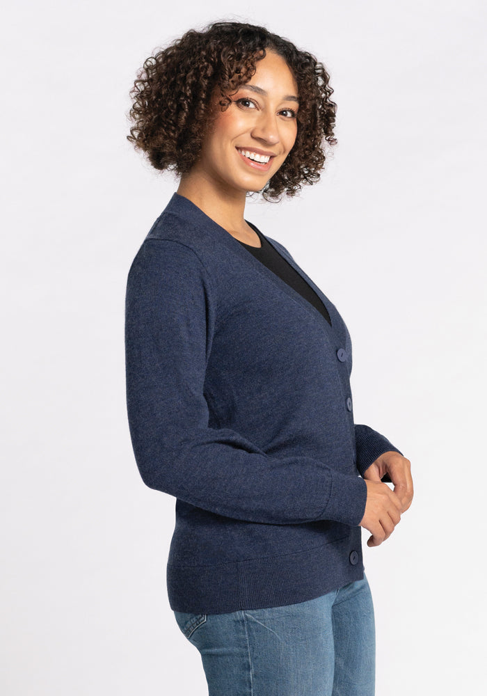 A person with curly hair is smiling and standing sideways to the camera. They are wearing a Woolx Gia Sweater in Twilight Storm over a black top and jeans, against a plain white background.