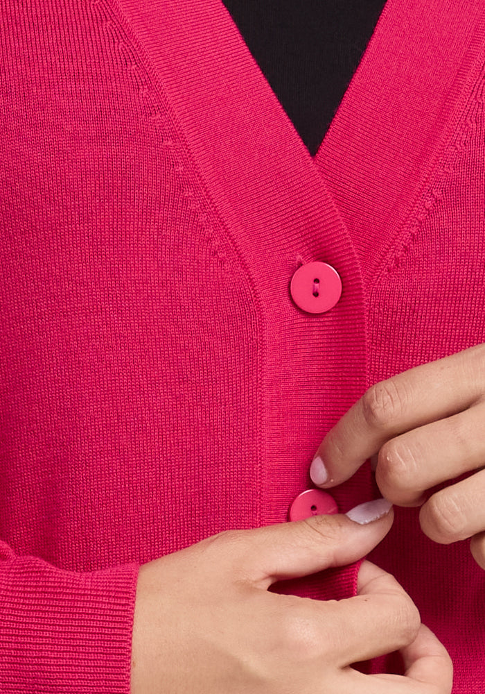 A person is buttoning a Woolx Gia Sweater in Berry Burst. The focus is on the hands and buttons, with a black shirt visible underneath. Crafted from soft, finely knit Merino wool, this piece offers lightweight warmth and luxurious comfort.