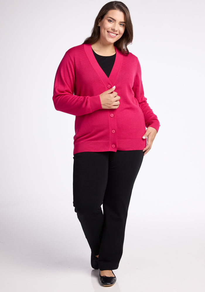 A person smiles while wearing the Woolx Gia Sweater in vibrant Berry Burst over a black top and pants. With one hand on the Merino wool sweater, they relish its lightweight warmth, standing against a plain white background.