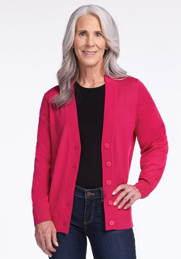 A woman with long gray hair smiles while wearing the Gia Sweater in Berry Burst from Woolx, paired with a black top and dark jeans. She stands against a plain white background.