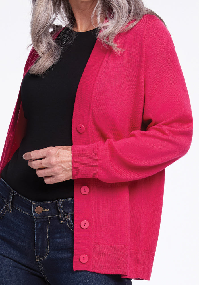 A person wearing the Woolx Gia Sweater in Berry Burst with large buttons, paired with a black top and dark blue jeans, stands against a plain white background. Their hair is long and gray.