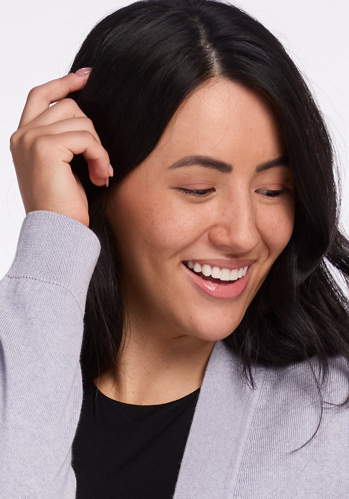 A person with long dark hair smiles while looking to the side, wearing a black top and the travel-ready Gia Sweater in Mist by Woolx. Their hand gently touches their hair, accentuated by the soft texture of Merino wool, against a white background.