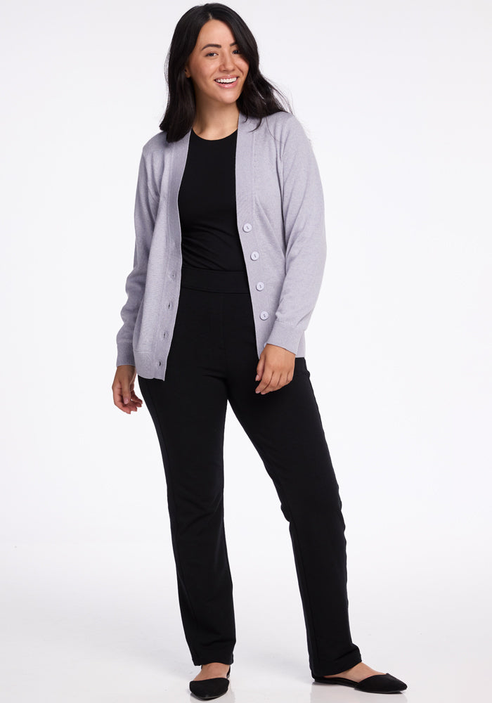 A woman with long dark hair smiles while wearing the Gia Sweater - Mist, a lightweight gray cardigan by Woolx made of Merino wool, over a black shirt and pants. She stands with her left hand on her hip and dons black flats, exuding a travel-ready vibe against the plain white background.