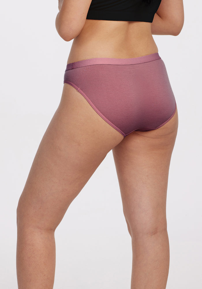 A person is shown from behind wearing Woolx Roxie Bikini Underwear in Wild Ginger and a black top against a plain gray background, highlighting the effortless stay-put comfort of the underwear.