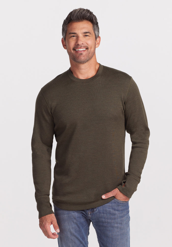 A man with salt-and-pepper hair smiles while wearing a dark green Woolx Glacier Long Sleeve Crew in the "Bark" color and blue jeans, standing against a plain white background with one hand in his pocket. 