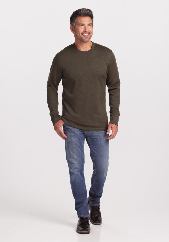 A man with short hair is walking and smiling, wearing the Woolx Glacier Long Sleeve Crew in Bark, along with blue jeans and black shoes. The background is plain white.