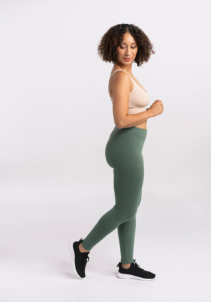 A person with curly hair is standing in a side profile pose against a plain white background. They are wearing a beige sports bra, Woolx Stella Leggings in Duck Green, and black athletic shoes. Their head is slightly turned and one foot is lifted slightly off the ground.