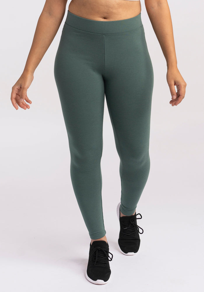 A person is wearing Woolx Stella Leggings in dark green high-waisted thermal design and black sneakers, perfect for a stylish winter wardrobe. The background is plain white, and the person is standing with their arms slightly away from their sides, showcasing only the lower half of their body. 