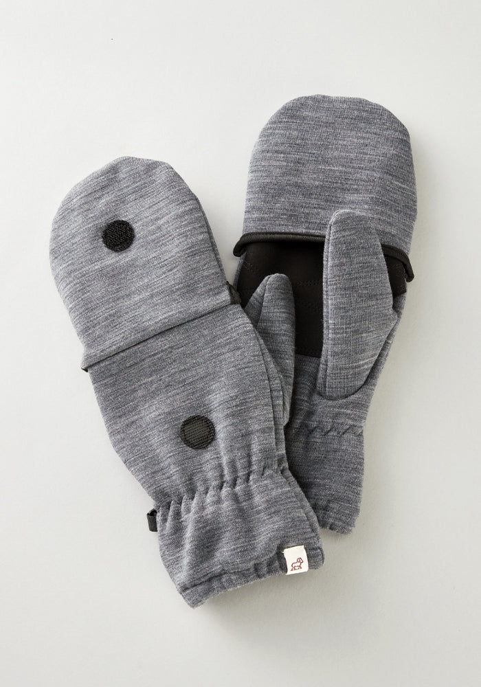 The Cameron Glove Mitten in Graphite Heather by Woolx boasts a versatile convertible design with elastic cuffs. Crafted from a blend of merino wool, these mittens feature black accents including a patch on the back and thumb area. A small tag adorned with a bear logo completes the look as they rest on a light grey surface.