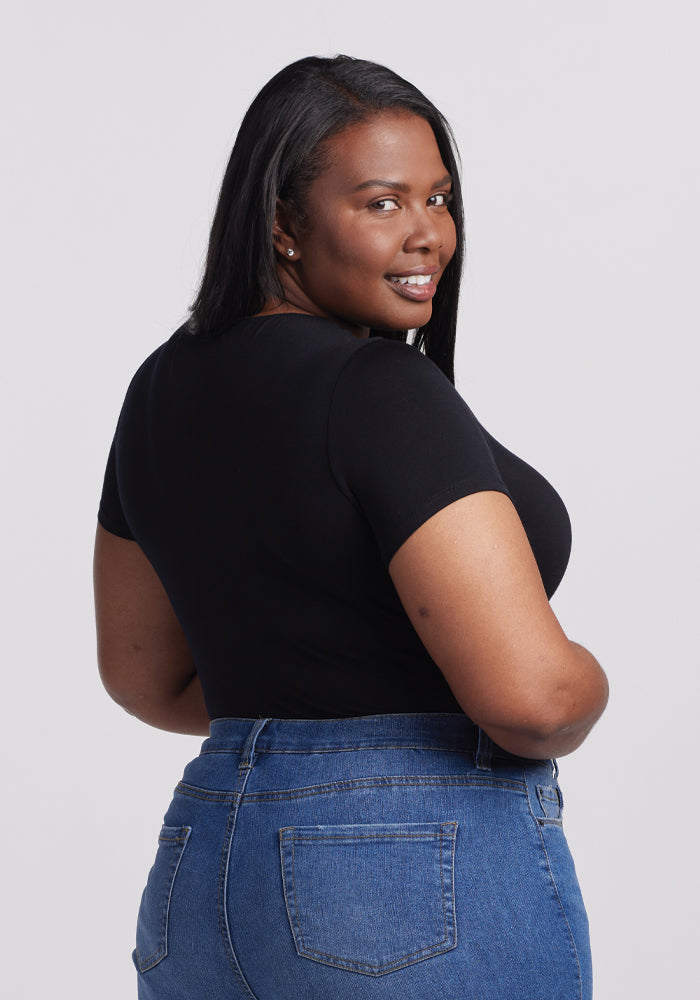 A woman with long dark hair smiles over her shoulder, wearing the sleek Gracie Bodysuit in black by Woolx and blue jeans, standing out against a plain light gray background.