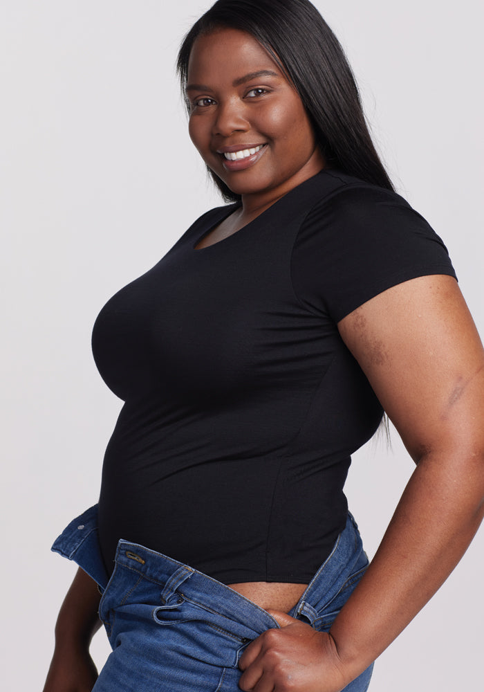 A woman smiles at the camera in a black Gracie Bodysuit by Woolx. She holds open the waistband of her blue jeans, with her long, straight black hair elegantly cascading as she stands against a plain white background.