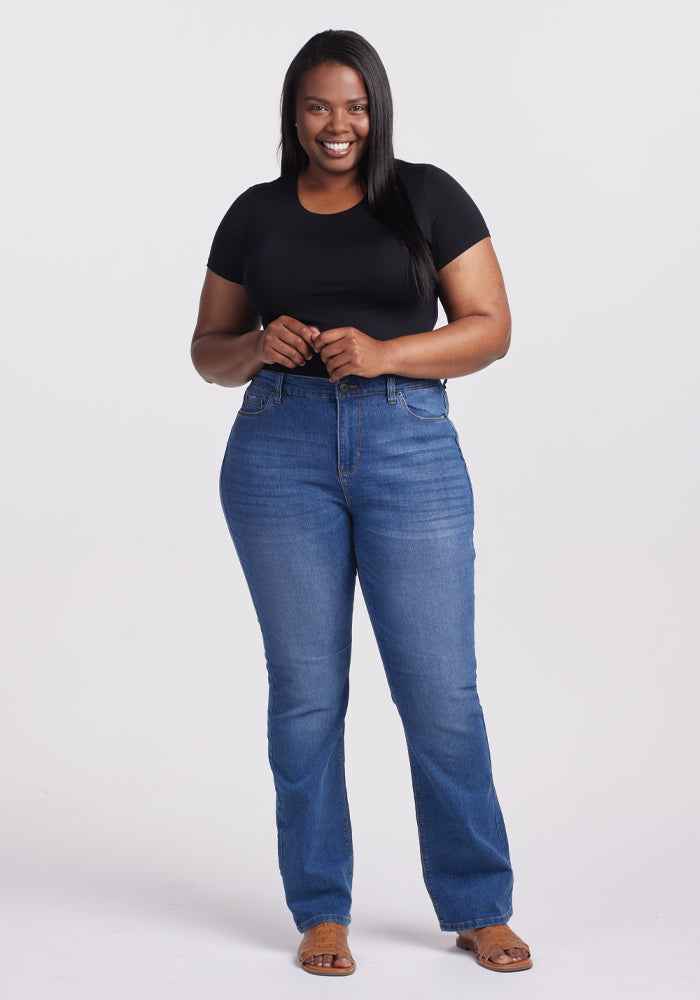 A woman with long black hair smiles while standing against a plain white background, wearing a Woolx Gracie Bodysuit in black, paired with blue jeans and sandals.