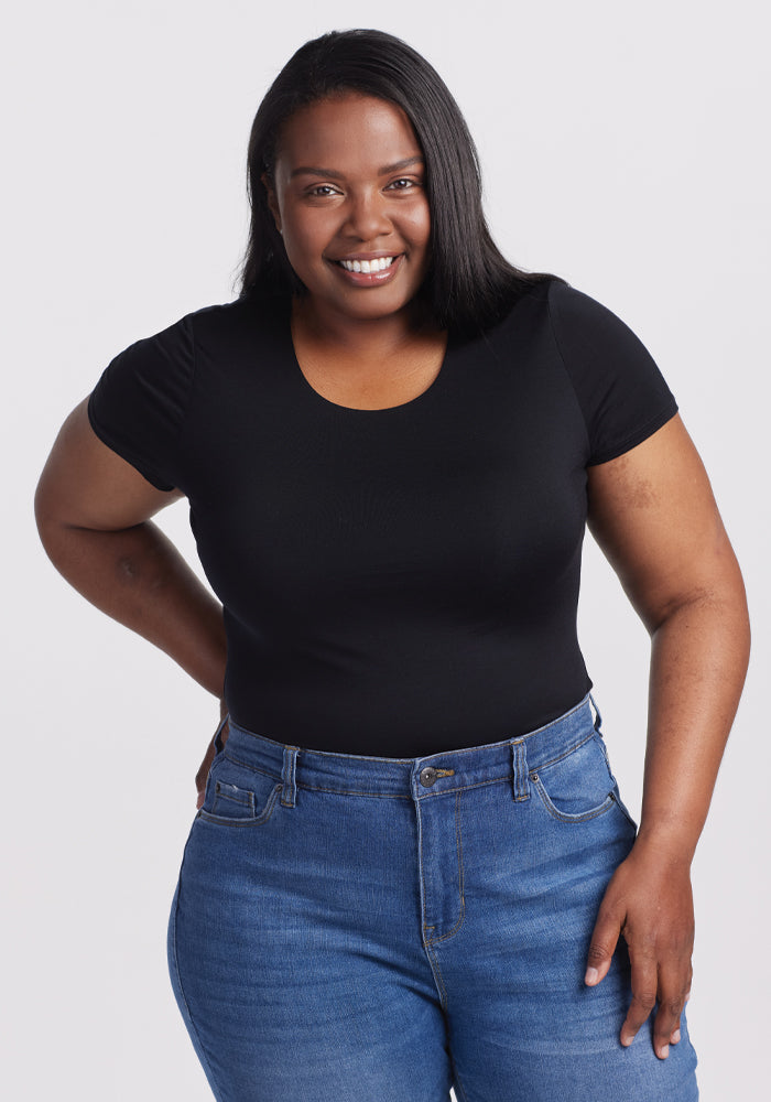 A smiling woman with long dark hair stands against a light gray background, showcasing the Woolx Gracie Bodysuit in black, seamlessly paired with blue jeans. Her hand rests confidently on her hip, emphasizing the elegance of her outfit. 