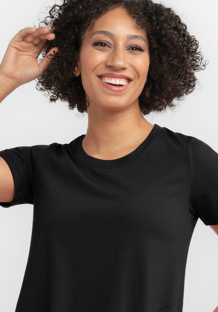 A person with curly hair is smiling while wearing Woolx's black Georgie Dress against a plain background, their hand raised to their head.