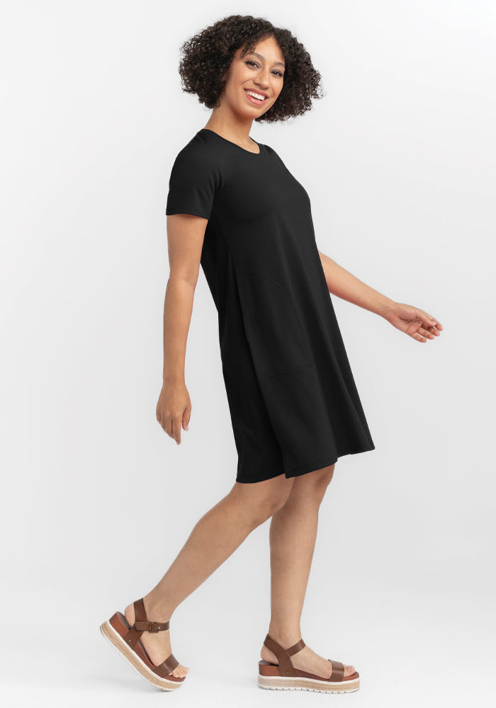 A person with curly hair smiles while moving sideways, wearing a short-sleeved black Georgie Dress by Woolx and brown platform sandals. The white background highlights their confident and relaxed demeanor. 