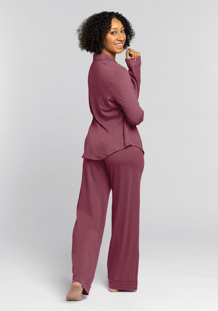 A person with curly hair stands barefoot, facing away and turning slightly towards the camera. They wear the comfortable Harper FeatherTouch® Pajama Set in Wild Ginger by Woolx, smiling against a plain grey background.