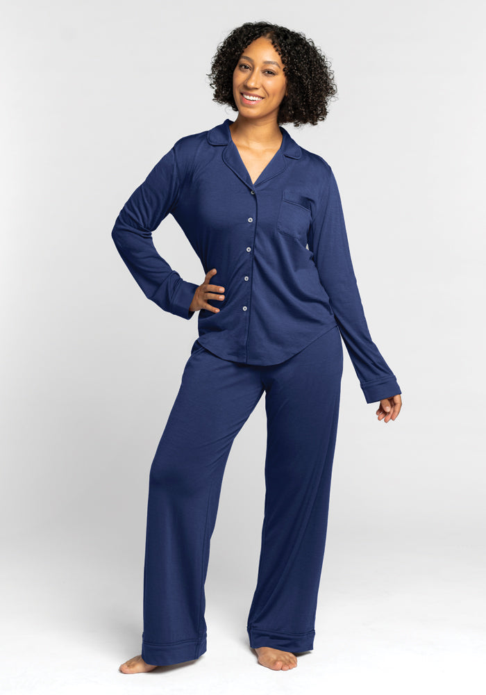 A person with curly hair is smiling and posing in a relaxed manner, wearing Woolx's Harper FeatherTouch® Pajama Set in Starry Night, complete with long sleeves and buttons. This relaxed fit pajama set perfectly complements their carefree vibe as they stand barefoot against a plain light gray background.