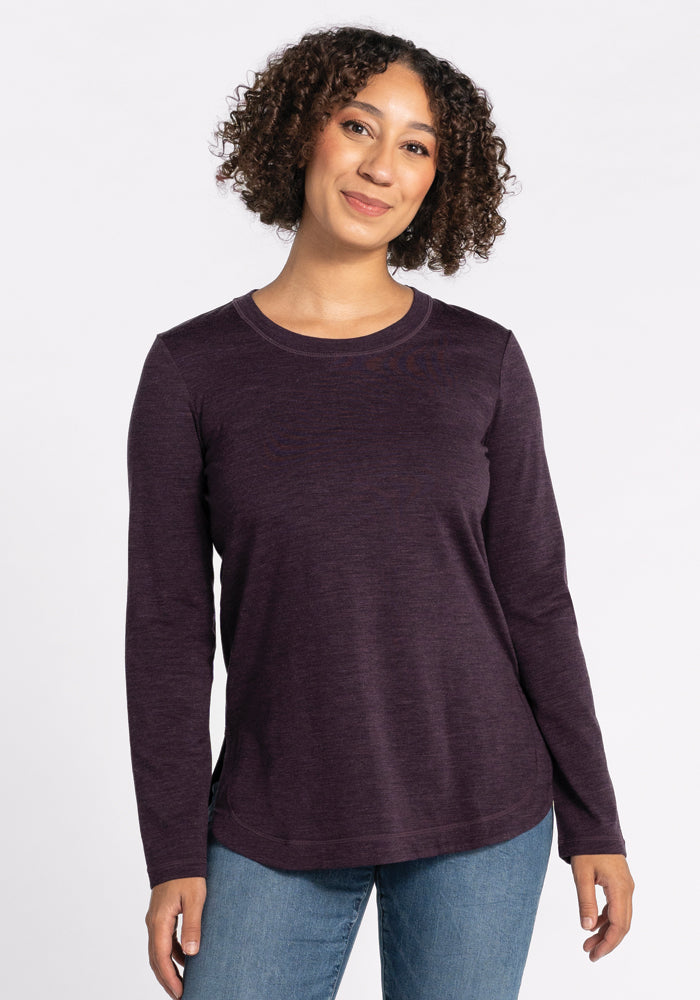 A person with curly hair is smiling while wearing a relaxed-fit, long-sleeved Hazel Tunic in Deep Plum from Woolx and blue jeans. The background is plain white. 