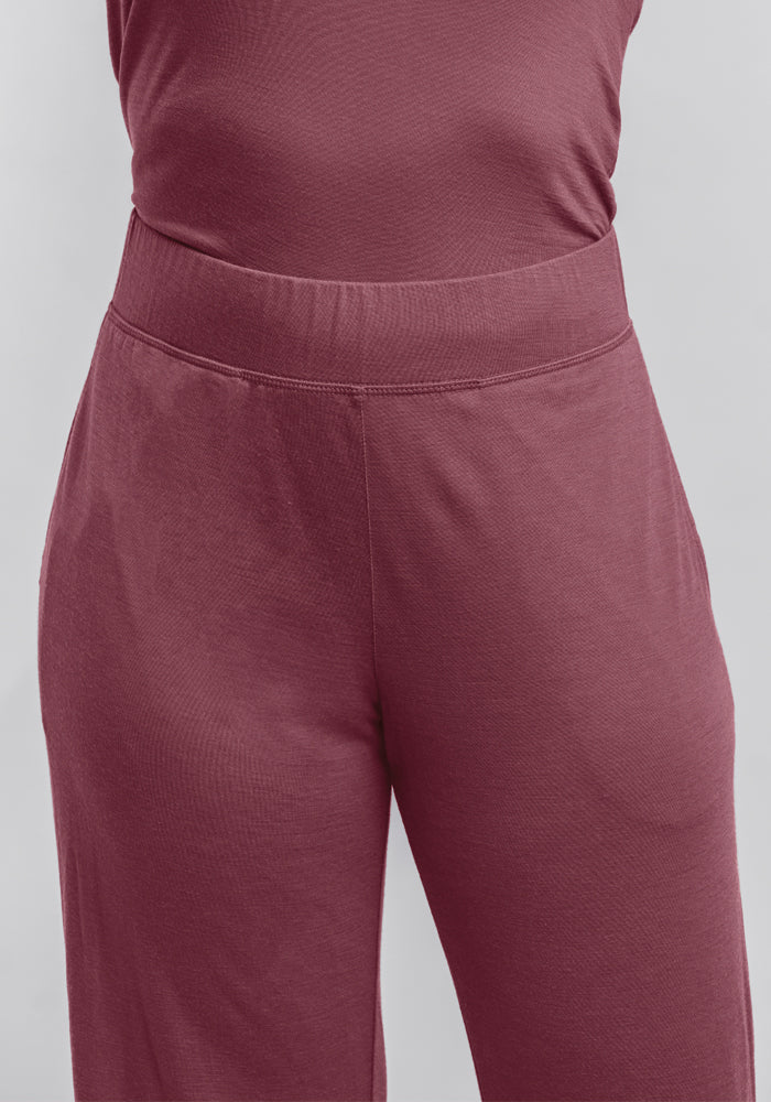 Close-up of a person wearing the Harper FeatherTouch® Pajama Set in Wild Ginger, from the brand Woolx. The pajama pants are maroon, high-waisted, and made from a soft, stretchy material akin to cozy PJs. Featuring a wide waistband and a smooth, wrinkle-free finish, these pants highlight an effortlessly comfortable and relaxed fit. The background features a plain light gray color.