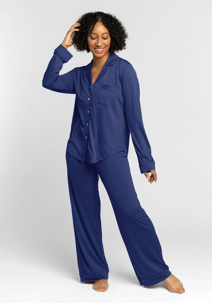 A person wearing the Woolx Harper FeatherTouch® Pajama Set in the Starry Night design smiles and poses against a plain white background. The relaxed fit set includes a button-up long-sleeve top and loose-fitting pants. They have curly hair and are barefoot. 