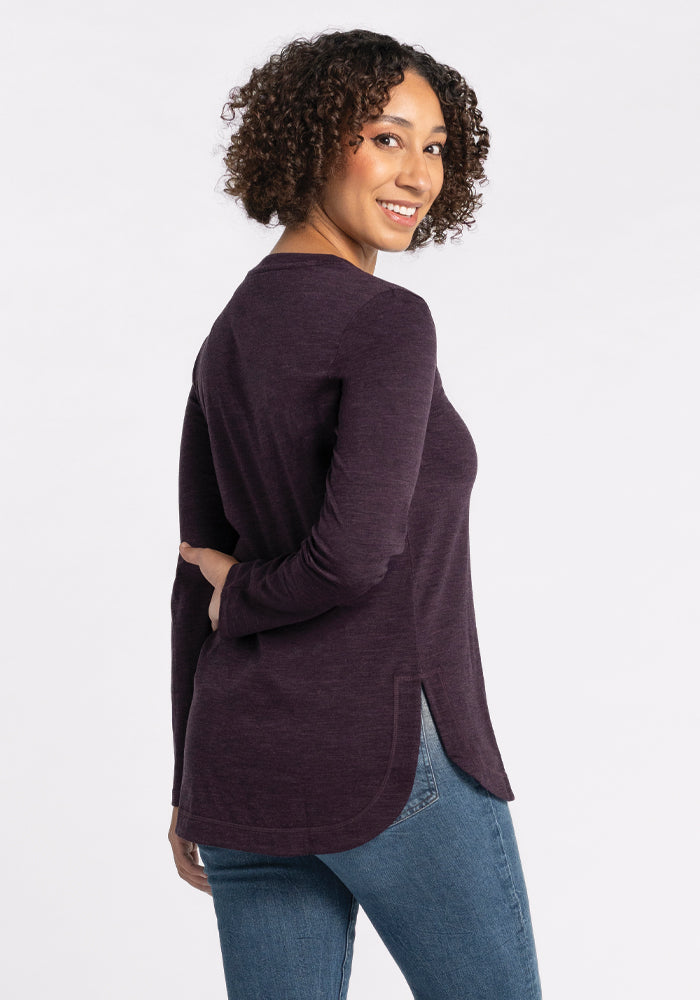 A person with curly hair smiles while looking over their shoulder, wearing the Hazel Tunic - Deep Plum by Woolx, crafted from merino wool in a relaxed fit. They pair it with blue jeans and stand against a plain white background.