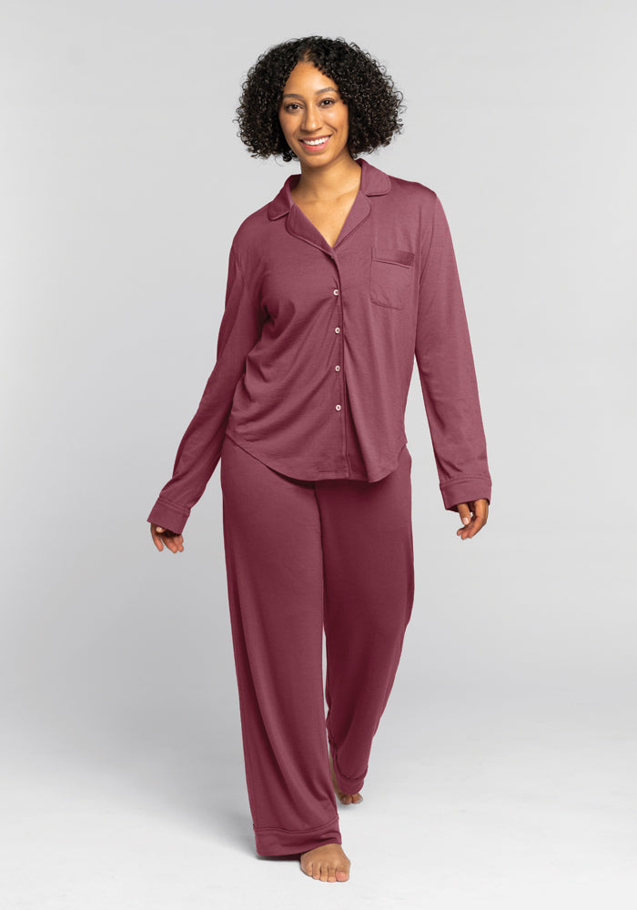 A person with curly hair is smiling and walking barefoot, wearing a cozy Woolx Harper FeatherTouch® Pajama Set in Wild Ginger, which consists of a long-sleeve shirt with buttons and loose-fitting pants. The background is plain gray.