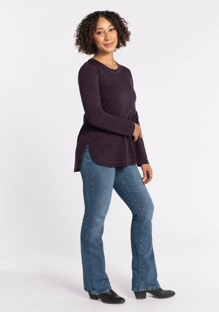 A person with curly hair is smiling, standing in a relaxed pose. They are wearing a stylish Hazel Tunic - Deep Plum from Woolx, paired with blue jeans. The background is plain white.