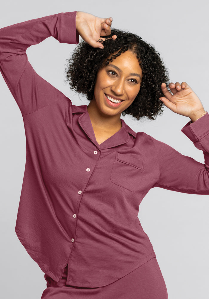 A person in a cozy Wild Ginger long-sleeve button-up shirt from Woolx poses with arms playfully raised. They have curly hair and a joyful expression, standing against a plain light gray background—like they've just slipped out of the Harper FeatherTouch® Pajama Set after a blissful sleep.