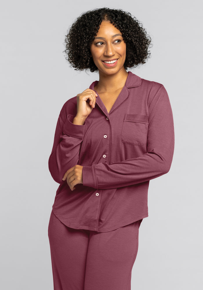 A curly-haired person smiles in a Woolx Harper FeatherTouch® Pajama Set in Wild Ginger. This burgundy travel set is made from temperature-regulating fabric and includes a collared button-up top with a pocket and long sleeves against a plain gray background.