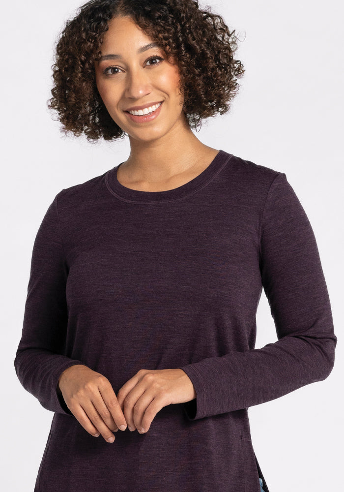 A person with curly hair smiles while wearing the Woolx relaxed fit Hazel Tunic - Deep Plum in long sleeves. The background is plain and light-colored.