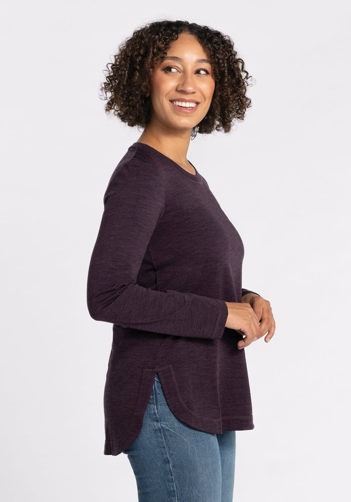 A person with curly hair smiles while gazing to the side, wearing the relaxed-fit Hazel Tunic in Deep Plum by Woolx and jeans, against a plain white backdrop.