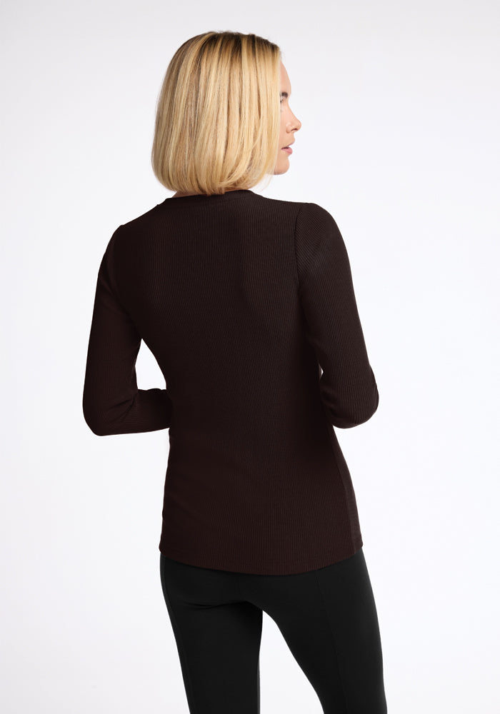 A person with shoulder-length blonde hair stands with their back to the camera, wearing a long-sleeved Woolx Hadley Ribbed Crew in French Roast and black pants. The background is plain and white.