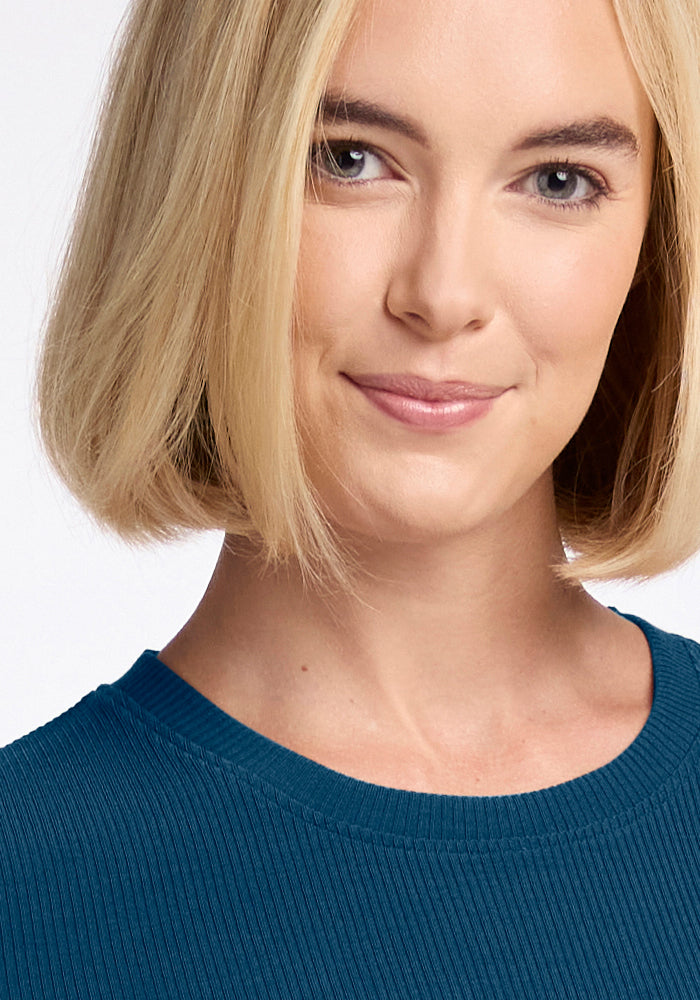 A person with short blonde hair is softly smiling at the camera, wearing a Woolx Hadley Ribbed Crew in Real Teal. The background is plain and light-colored.