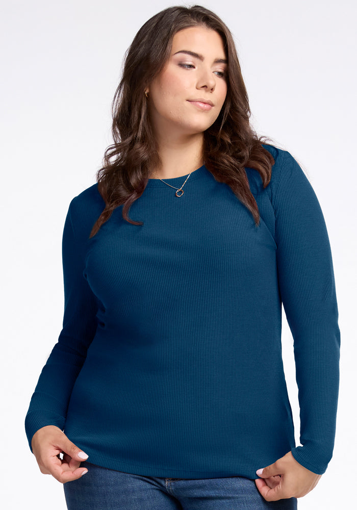 A person with long dark hair is wearing the Woolx Hadley Ribbed Crew in Real Teal along with jeans. They are looking to the side, standing against a plain white background. 