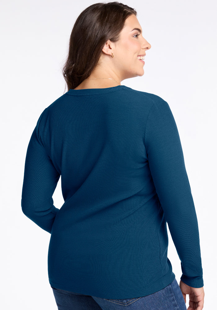 A person modeling the Woolx Hadley Ribbed Crew in Real Teal is depicted from the side and back. They have long brown hair and are smiling with their arms relaxed, set against a plain white background.