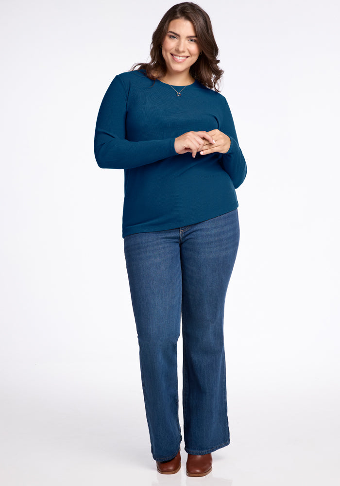 A person with long dark hair smiles while standing, dressed in a Real Teal Hadley Ribbed Crew top by Woolx, perfectly paired with blue jeans and brown shoes. The simple white backdrop accentuates the elegance of their outfit made from Australian Merino Wool.