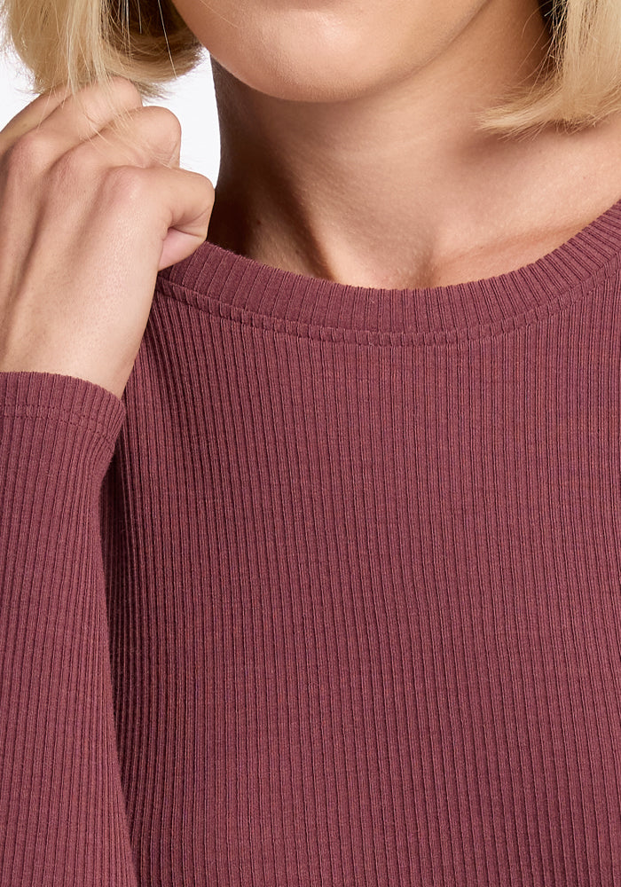 A close-up image of a person with short blonde hair wearing a Woolx Hadley Ribbed Crew in Wild Ginger. Their hand gently touches the crew neckline, emphasizing the texture of Merino Wool and the elegant positioning of their fingers.