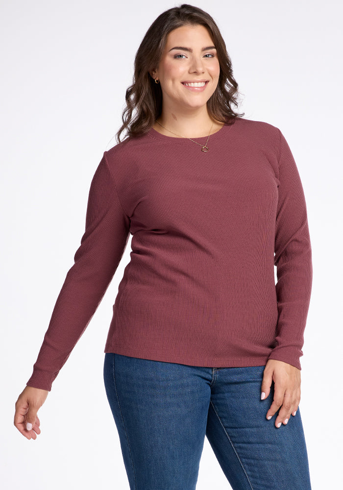 A woman with long brown hair smiles at the camera, wearing the Hadley Ribbed Crew in Wild Ginger by Woolx, paired with blue jeans. She stands with one hand slightly raised and the other resting at her side against a plain white background.