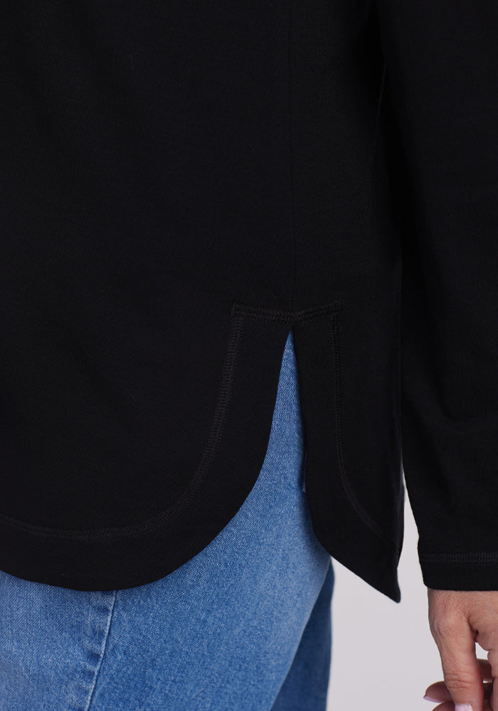 A close-up photo showcases a person wearing the black, lightweight Hazel Tunic by Woolx, featuring Merino wool with temperature-regulating fabric, rounded hem, and side slits. The tunic is paired with blue jeans, highlighting its lower part and one casually hanging arm.