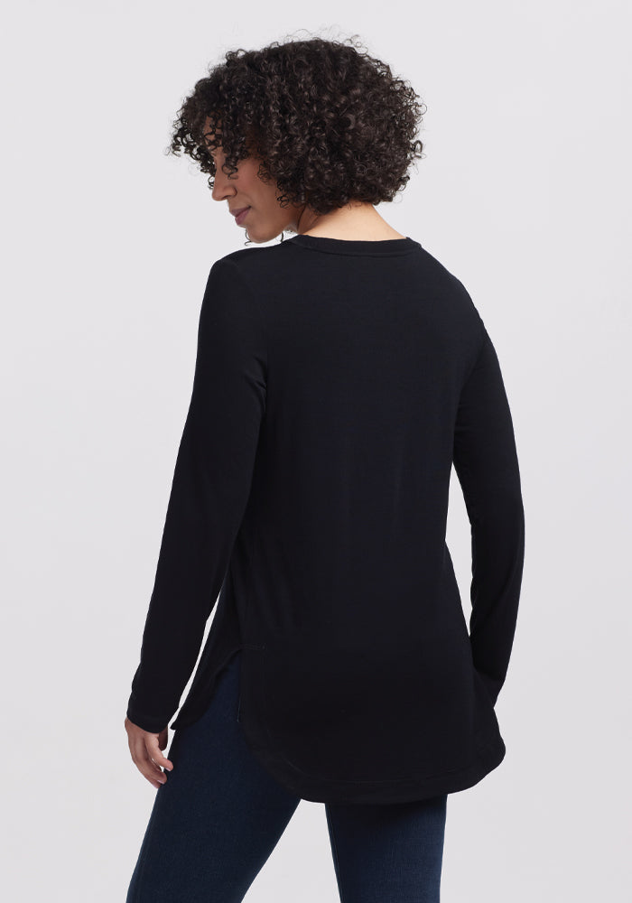 A person with curly hair, turned away, wears the Woolx Hazel Tunic in black and jeans. The plain white background underscores their simple style.