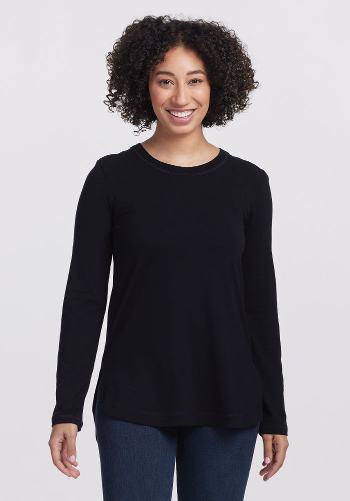 A person with curly hair smiles in the Woolx Hazel Tunic—an elegant long-sleeve black shirt made of temperature-regulating fabric. They pair it with jeans, standing against a plain white background. 