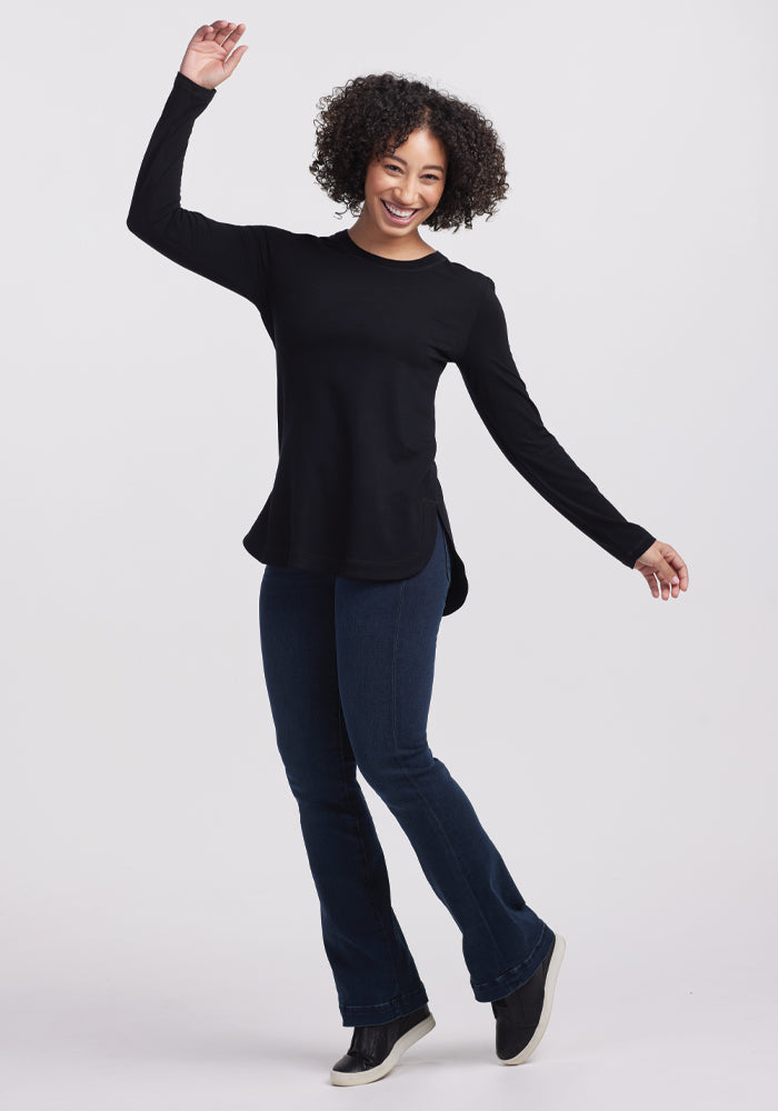 A person with curly hair playfully poses in a lightweight Woolx Hazel Tunic - Black, paired with dark jeans and black shoes against a plain white background, showcasing an effortlessly chic and temperature-regulating outfit.