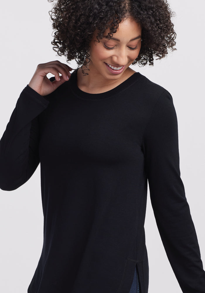 A curly-haired person smiles while wearing the Woolx Hazel Tunic in Black, standing against a plain light background. The long-sleeved Merino wool tunic drapes gracefully around them, exuding comfort and style with its temperature-regulating, lightweight fabric.