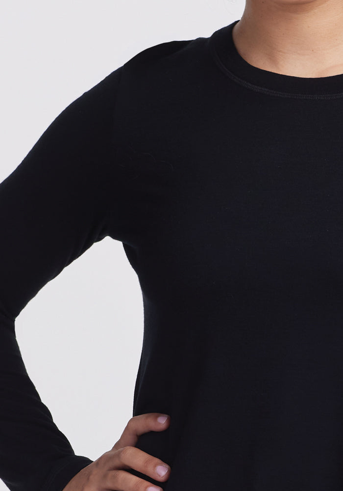 A person wears Woolx's Hazel Tunic in black, a lightweight merino wool long-sleeve shirt. The image focuses on the torso, emphasizing the tunic's texture, fit, and temperature-regulating fabric against a plain light background as they stand with their right hand on their hip.