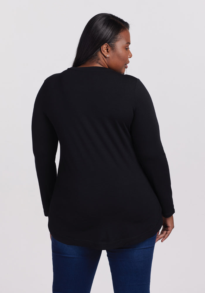 The individual is facing away from the camera, displaying the Woolx Hazel Tunic in black, a temperature-regulating long-sleeved top styled as a lightweight tunic over blue jeans. The background is plain and light-colored.