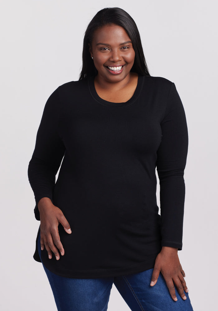 A person with long hair smiles confidently, wearing a lightweight Woolx Hazel Tunic in black and blue jeans. They stand against a plain white backdrop, one hand on their hip.