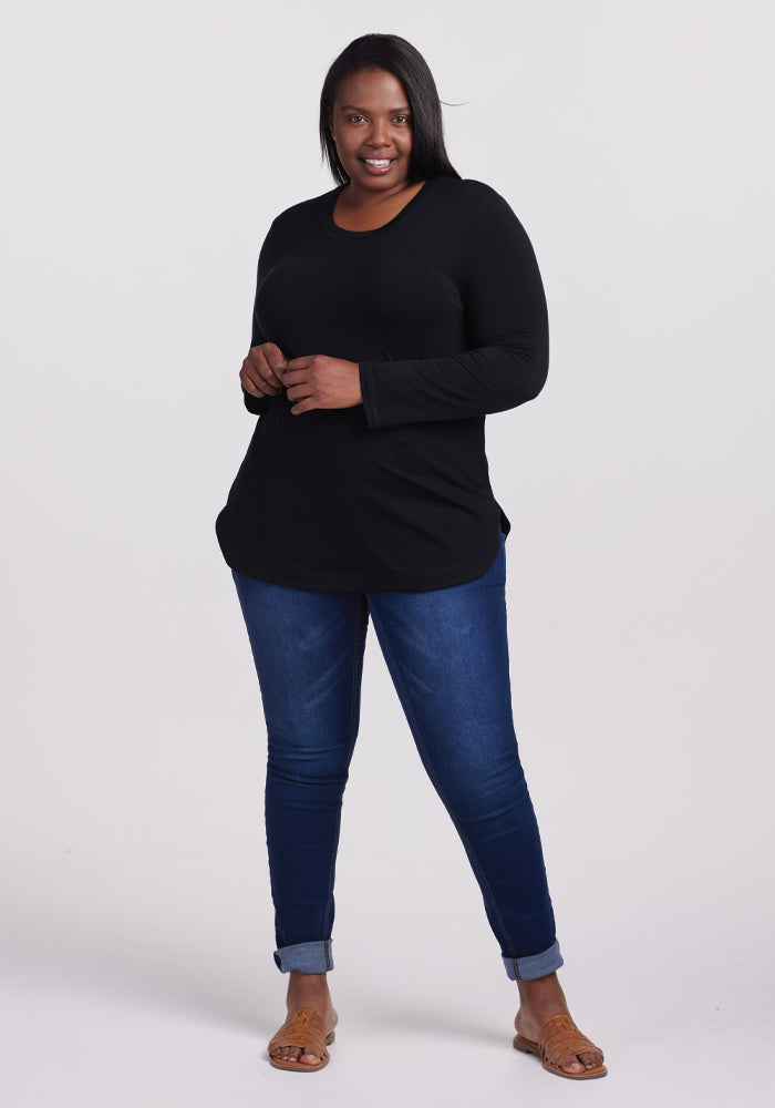 Clad in a Woolx Hazel Tunic in black and dark blue jeans, the confident figure smiles with long, dark hair. The Merino wool tunic offers temperature-regulating comfort. Sandals appear as the jeans roll slightly at the ankles against a plain white background.