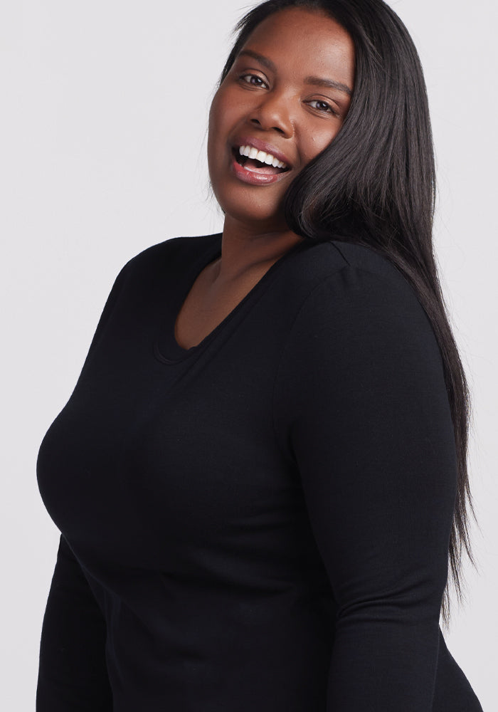 A person with long dark hair is smiling widely, dressed in a black Woolx Hazel Tunic against a plain light gray background.